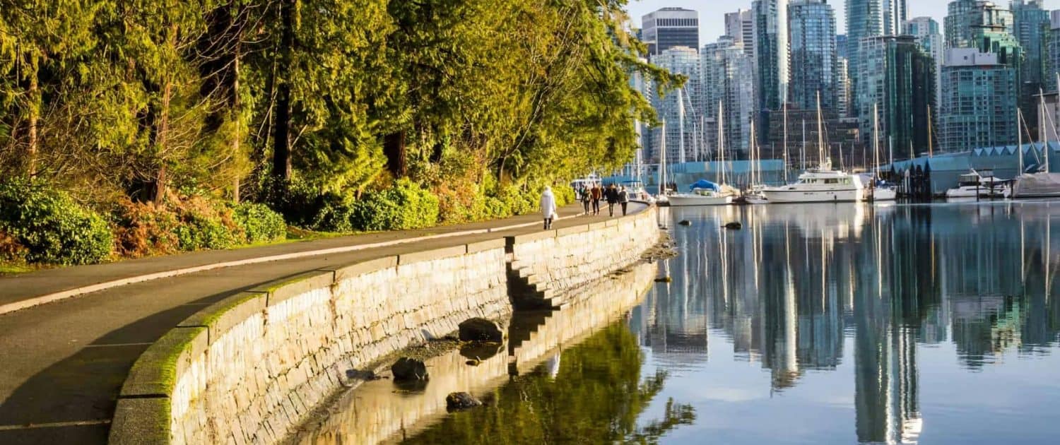 Trendy urban waterfront with lush greenery, modern skyscrapers, and sailboats reflecting on calm waters in downtown Vancouver, showcasing a blend of nature and city life.