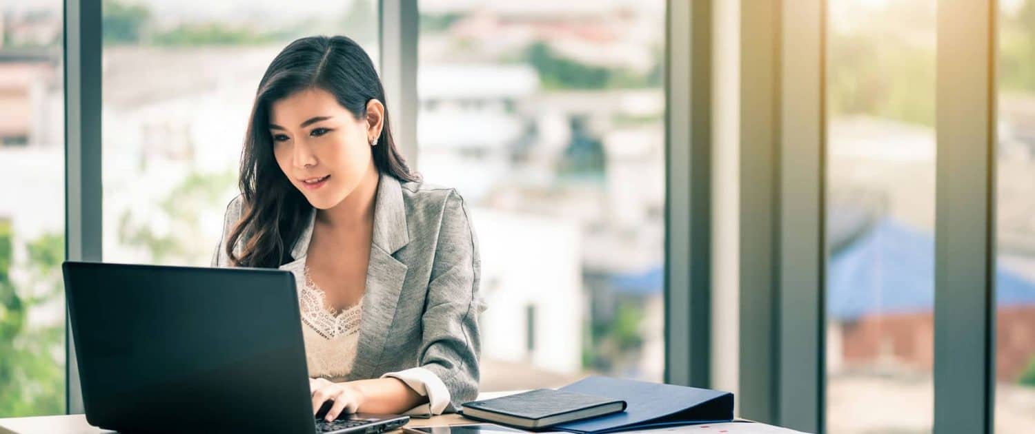 Professional woman working on a laptop in a modern office with large windows and city view.