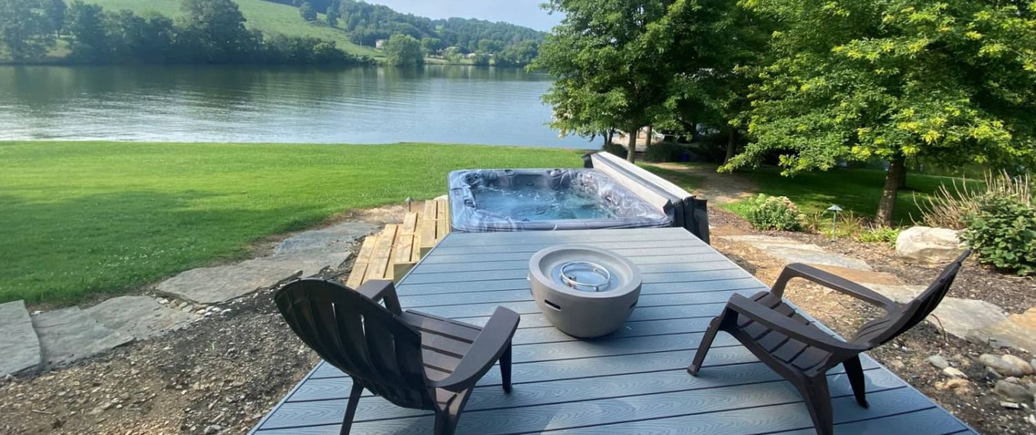 Relaxing lakeside patio with hot tub, two Adirondack chairs, and scenic water view, ideal for outdoor leisure and enjoying nature.